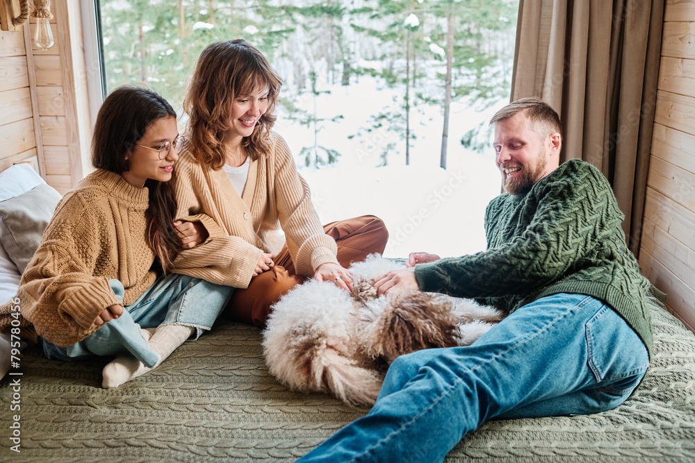 Caucasian man, woman and teen girl spending winter day indoors relaxing on bed with their dog in country house Caucasian man, woman and teen girl spending winter day indoors relaxing on bed with their dog in country house