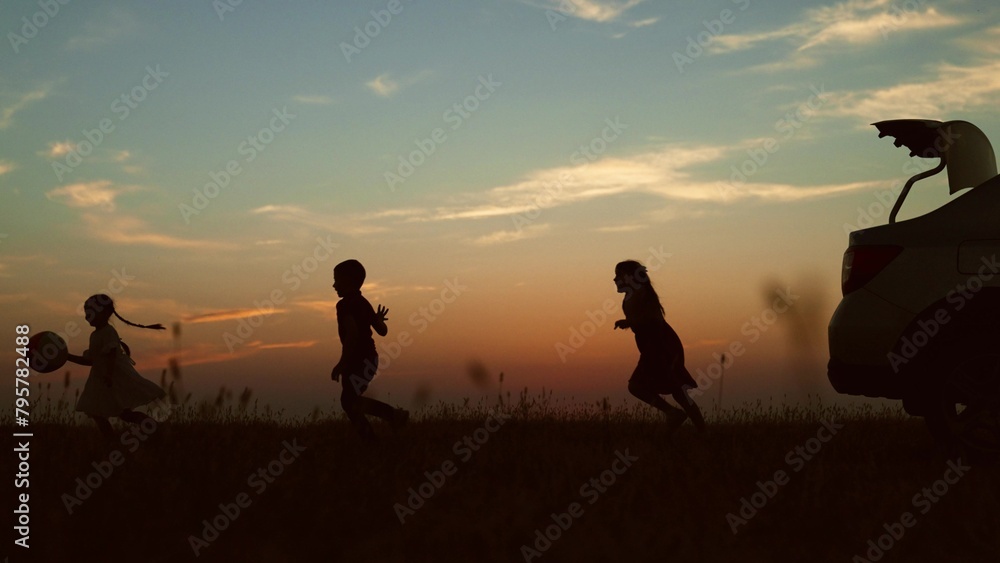 Silhouettes of children playing at sunset by car. Cute cheerful funny kids children siblings brothers sisters run with ball play catch-up in field park nature. Close-knit family on journey travel trip