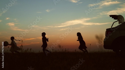 Silhouettes of children playing at sunset by car. Cute cheerful funny kids children siblings brothers sisters run with ball play catch-up in field park nature. Close-knit family on journey travel trip