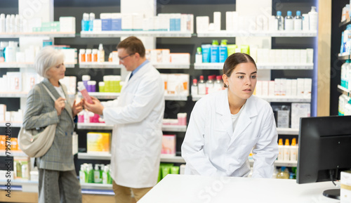 Attentive young female pharmacist looking at display of computer in chemist's shop with large assortment