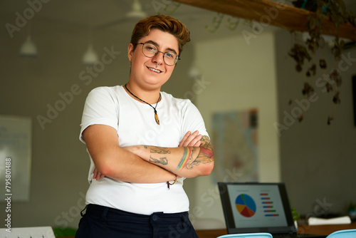 Confident transgender professional stands in modern office, arms crossed, smiling. Inclusive work environment, laptop with charts in background. Pride, equality themes in corporate setting. photo