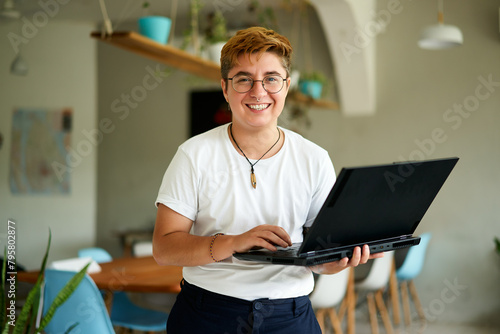 Cheerful transgender professional holds laptop, smiles in modern office. Gen Z using tech for work, represents inclusivity. Proud, confident posture, casual style, indoor plants background.