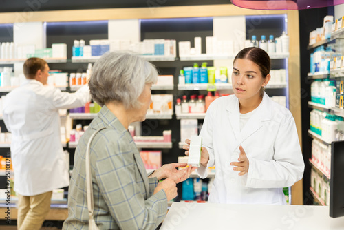Polite young female pharmacist consulting old costumer about care product in box in chemist s shop