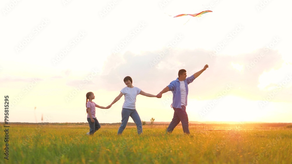 Happy family playing with flying kite at sunset summer field enjoy weekend leisure activity. Father mother and daughter holding hands walking at sunny meadow with flight toy unity and togetherness