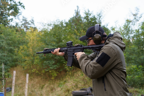 A man in a green jacket is holding a rifle and wearing headphones. He is in a forest and he is practicing shooting