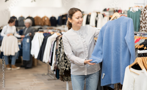 Happy middle-aged woman buyer choosing convenient jumper in clothing shop with large assortment