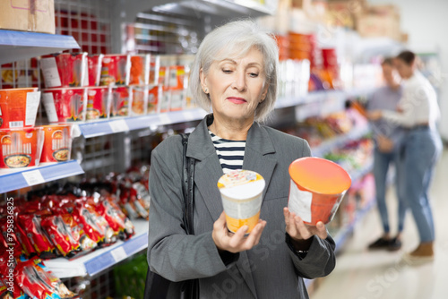 Senior woman in supermarket buys instant fast food in bowl. Mature female buyer reads information on packaging of semi-finished product. Concept of affordable box-container feeding
