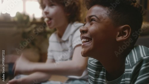 Side closeup of gen Y African American boy laughing so hard while watching movie with Caucasian best friend at home, nostalgic childhood in 90s