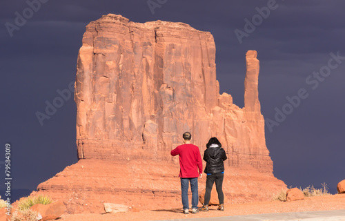 West Mitten, Monument Valley, illuminated by setting sun with very dark sky in background. Man and woman silhouetted in front.