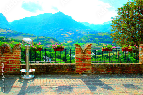 Tidy view from Montefortino at a town balcony and belvedere with brickwork merlons, metal barriers, vases of pink and green flora, panoramic binocular, a tree, hills, thickets, and the Sibillini