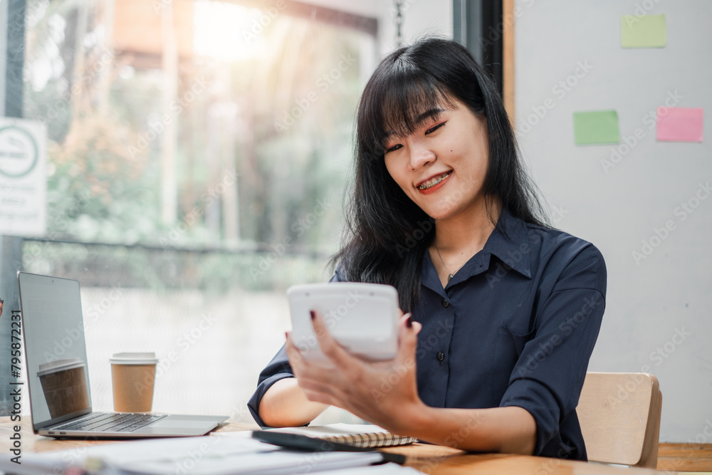 Smiling businesswoman with braces using calculator at her desk with a laptop and coffee in a bright workspace. Smiling businesswoman with braces using calculator at her desk with a laptop and coffee in a bright workspace.