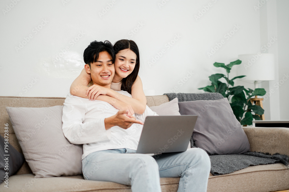 Young Asian couple enjoys a cozy moment on their sofa, interacting with a laptop, in a light-filled, plant-adorned living space. Young Asian couple enjoys a cozy moment on their sofa, interacting with a laptop, in a light-filled, plant-adorned living space.