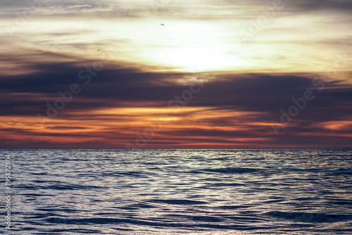 sunset over the sea nature background, backdrop, wallpaper, calm ocean ripples with light shining down from behind streaky or linear clouds in an orange sky and a distant horizon