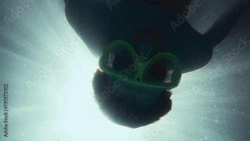 Male figure appearing through streaming sun light underwater to look into camera and rotate. Man having fun diving in swimming pool with mask and taking action camera. 