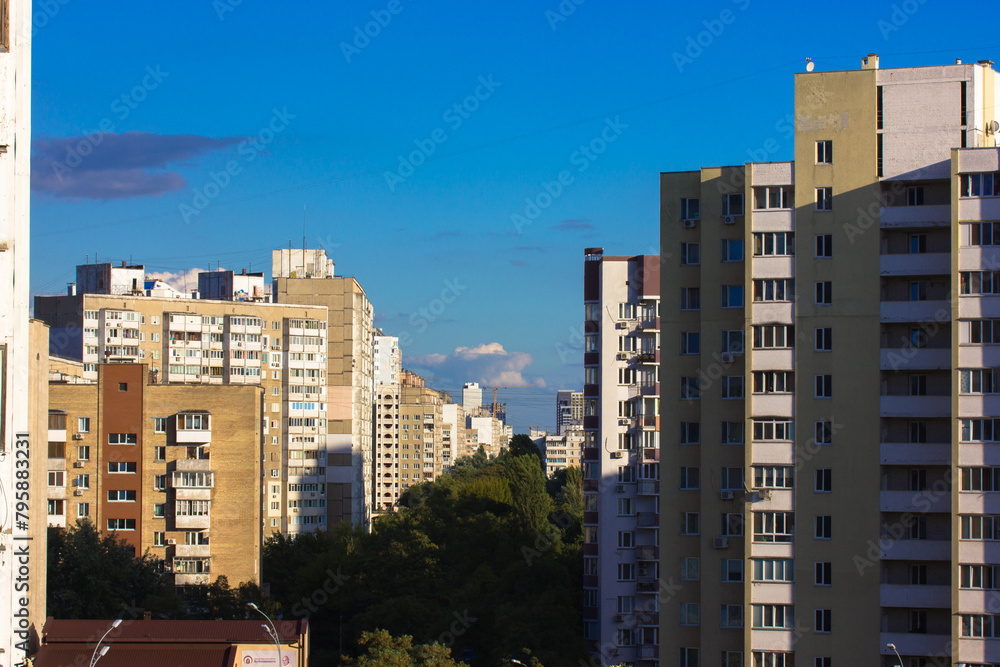 Multistory buildings against a blue sky at sunny day. A city street stretching into a distance buried in green trees. Walls of high post soviet union buildings. Ugly architecture. View from a window.
