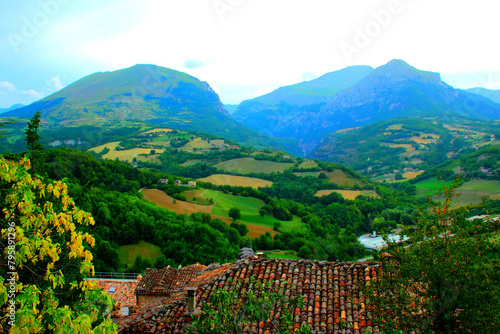 Balmy scene from Montefortino with yellow, green trees and brownish rooftops in the foreground, opening to suffused green hills punctuated by buildings and thickets, leading upwards to the Sibillini