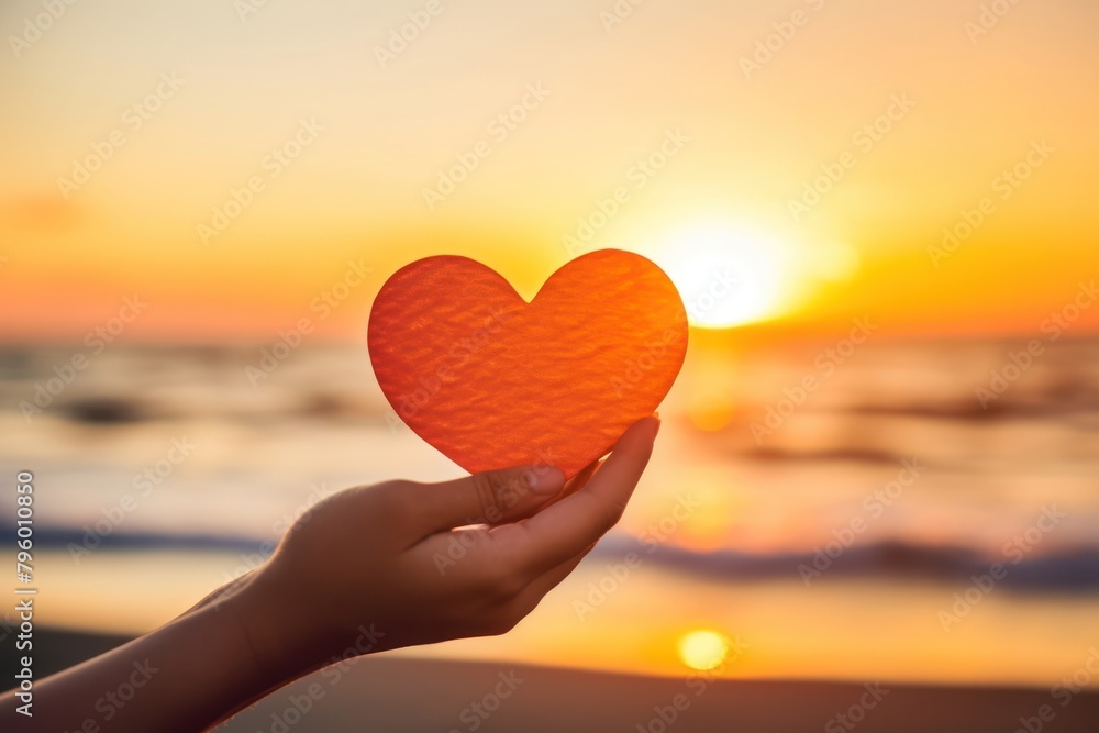 Fototapeta premium Hand holding a cut out of heart shaped paper against a sunset beach sunlight sky tranquility.