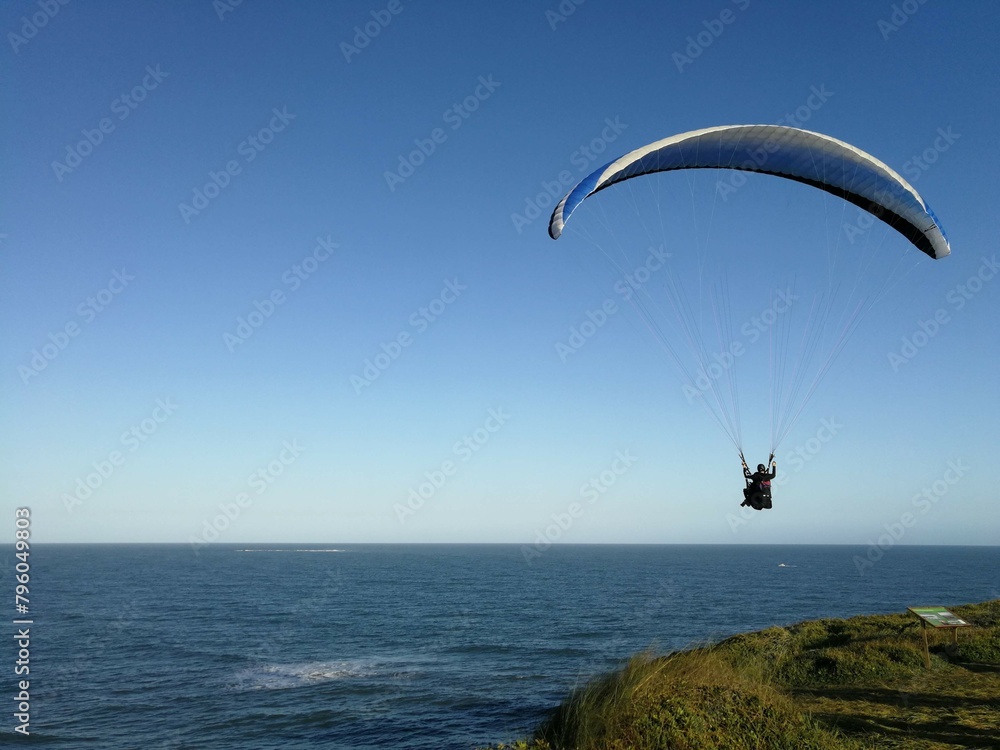 Pessoa voando de paraglider, com mar ao fundo, dia de céu azul, sem nuvens Pessoa voando de paraglider, com mar ao fundo, dia de céu azul, sem nuvens
