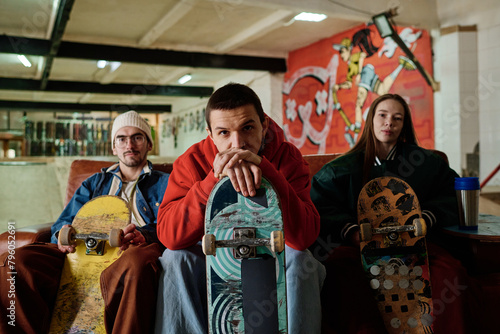 Self-confident young man and his male and female friends sitting on couch in skatepark posing for camera with skateboards, copy space