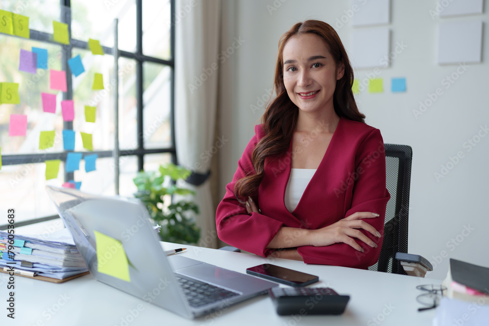 Confident Asian businesswoman with a smiling face sits with her arms crossed. Use a laptop to work and document analysis of financial reports. Office investment.