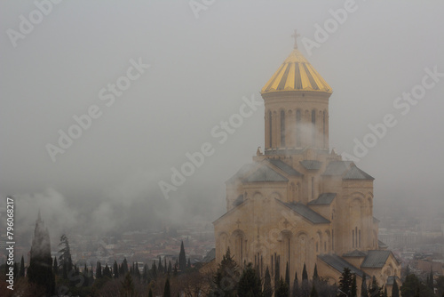 Tsminda Sameba in foggy weather in morning. Golden dome. View of Georgian Orthodox Church. Holy Trinity Cathedral of Tbilisi. Fog, smog. Cypress trees. Concept of faith in God, religion, tradition