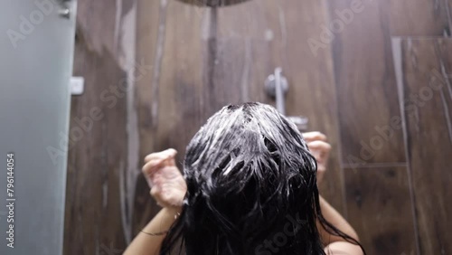 Rear view close up of a young Asian woman with long dark hair standing under the shower in a modern bathroom, washing and rinsing her hair, slow motion. photo