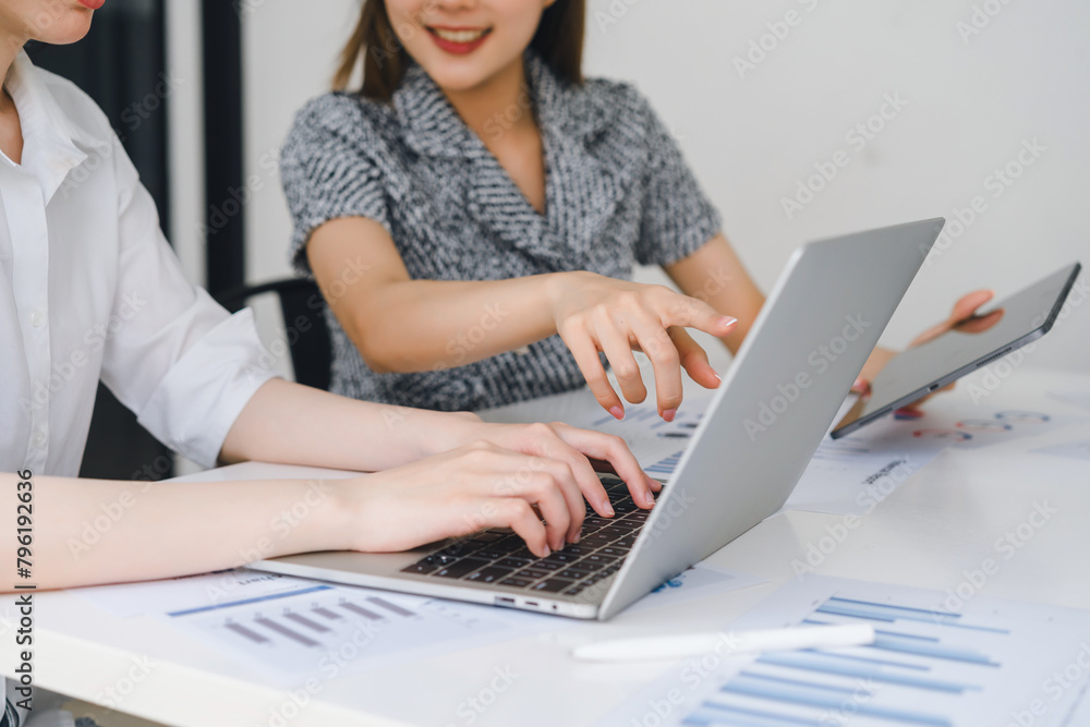 Two female colleagues working together on project, using laptop at a white desk with documents. Two female colleagues working together on project, using laptop at a white desk with documents.