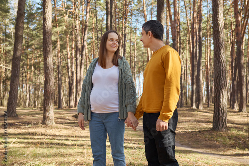 Brown haired pregnant woman walking in spring forest with her husband holding hands looking at each other with tenderness spending sunny days together