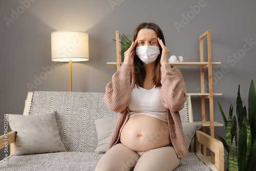 Sick pregnant woman wearing medical mask suffering terrible headache and flu symptoms massaging her temples while sitting on couch in cozy living room at home