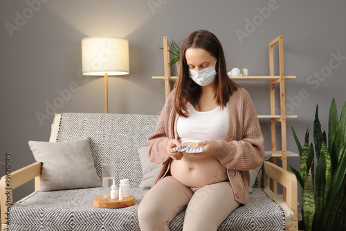 Unhealthy Caucasian brown haired pregnant woman wearing medical mask treating her grippe symptoms at home holding blister of pills treating her disease during pregnancy