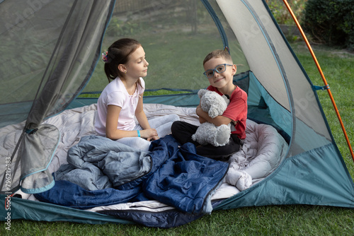 Two children, brother and sister, are sitting in a tent on a warm sunny day. Summer active entertainment, adventure and family recreation in nature. Children's camping. Healthy lifestyle