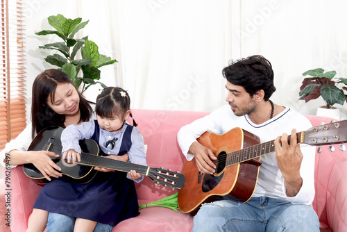 Happy Asian family. Chubby little girl daughter learning to play acoustic guitar with her dad. Child with mother and father spending time together in living room. Kid and parents have fun at home