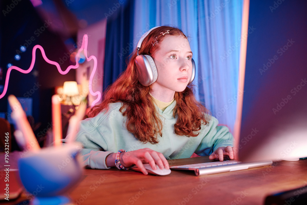 Medium shot of red-haired teenager sitting at desk in neon bedroom playing online video game on desktop computer Medium shot of red-haired teenager sitting at desk in neon bedroom playing online video game on desktop computer