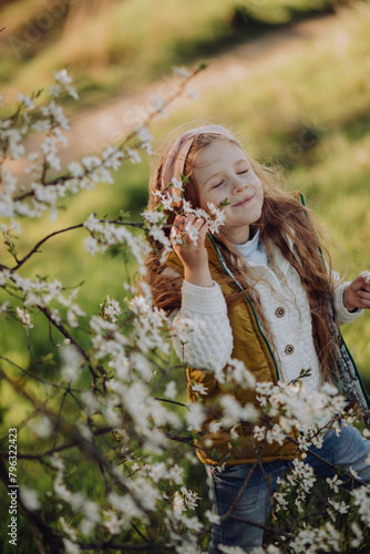little cute girl with long hair enjoying spring, admiring flowers on a blooming tree on a sunny day. smiling, enjoying the good weather. walk, family leisure, vertical portrait