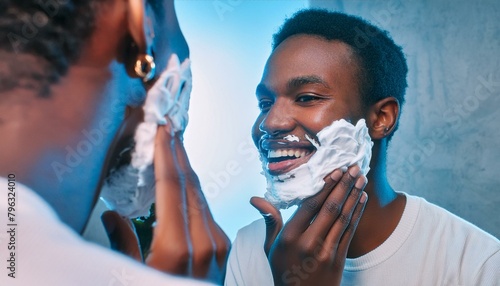 Close up over the shoulder of a young smiling black man shaving in the mirror.