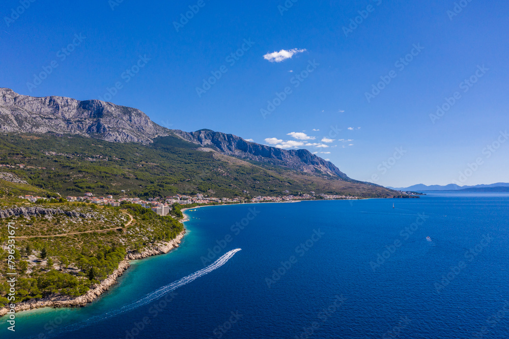 A small boat with tourists slowly floats on clear water. Amazing coastline. Aerial photography. The mountains are covered with greenery, the blue transparent sea. Makarska Riviera. Croatia