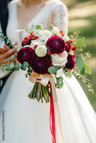 Bride in elegant dress holds vintage bouquet outdoors. Bouquet with white flowers roses, red dahlia, greens eucalyptus and plants, leaves. Wedding day. Beautiful bride portrait outdoors. Closeup.