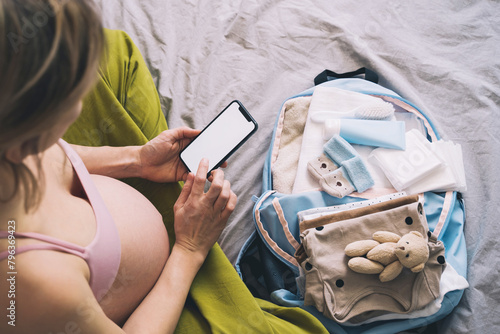 Pregnant woman getting ready for labor packing stuff for hospital, making notes or checklist in smartphone. Baby clothes, necessities for mother and newborn in maternity bag. photo