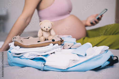 Baby clothes, necessities for mother and newborn in maternity bag. Pregnant woman getting ready for labor packing stuff for hospital, making notes or checklist in smartphone.