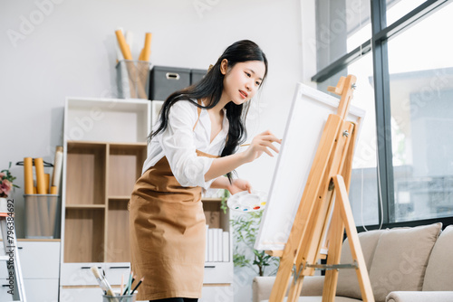 Asian Female painter do artwork in art workshop, painting supplies, oil pastels, two canvas easel, creative space with paintbrush in art studio