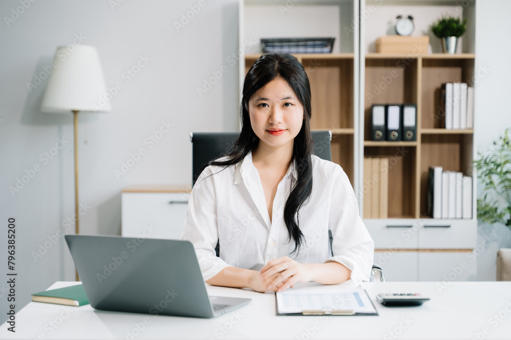 Young Asian woman smiling thinking planning writing in notebook, tablet and laptop looking at camera at office Young Asian woman smiling thinking planning writing in notebook, tablet and laptop looking at camera at office