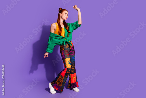 Full size photo of lovely woman dressed green shirt vintage pants dancing look at promo empty space isolated on purple color background