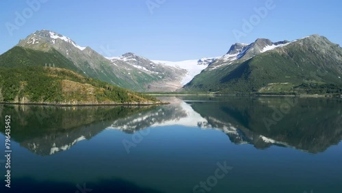 Drone slowly approaches the Svartisen Glacier, capturing a majestic landscape. The glacier sparkles in the sunlight, and the surrounding mountains reflect in the calm waters of the fjord photo