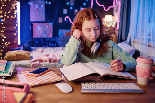 Medium shot of Caucasian girl with red hair sitting at desk in her room doing school homework in evening  copy space