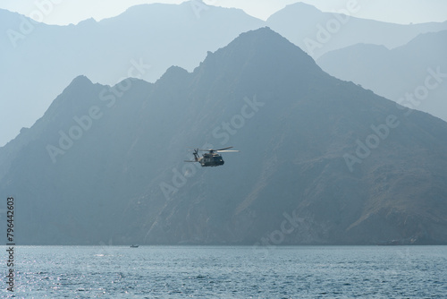 The helicopter soars gracefully over the calm waters while the mountains of Oman provide a breathtaking backdrop, capturing the essence of adventure and natural beauty. photo