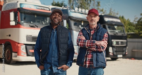Two mixed-race coworkers standing before large trucks outside. Mature drivers in hats and jackets positively looking into camera. Getting ready to traveling. Working together in delivery company.