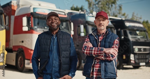 Two mixed-race coworkers standing before large trucks outside. Mature drivers in hats and jackets positively looking into camera. Getting ready to traveling. Working together in delivery company. photo