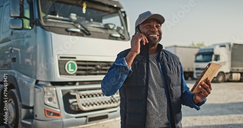 African American man speaking on phone with his coworker or customer while holding tablet device. Busy worker standing at trucks parking while remotely communicating. Concept of technology. photo