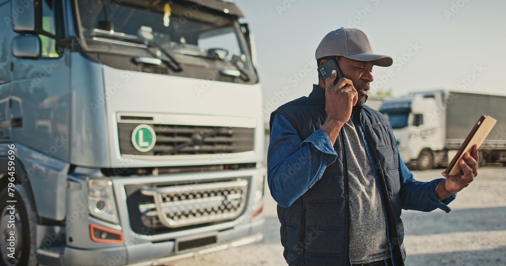 Portrait of african american worker talking by phone in background parked truck vehicles. Truck driver holding tablet, and checking route for new destination. Transportation service.