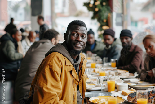 Positive homeless black man standing at the table in a street dining hall, surrounded by other individuals photo